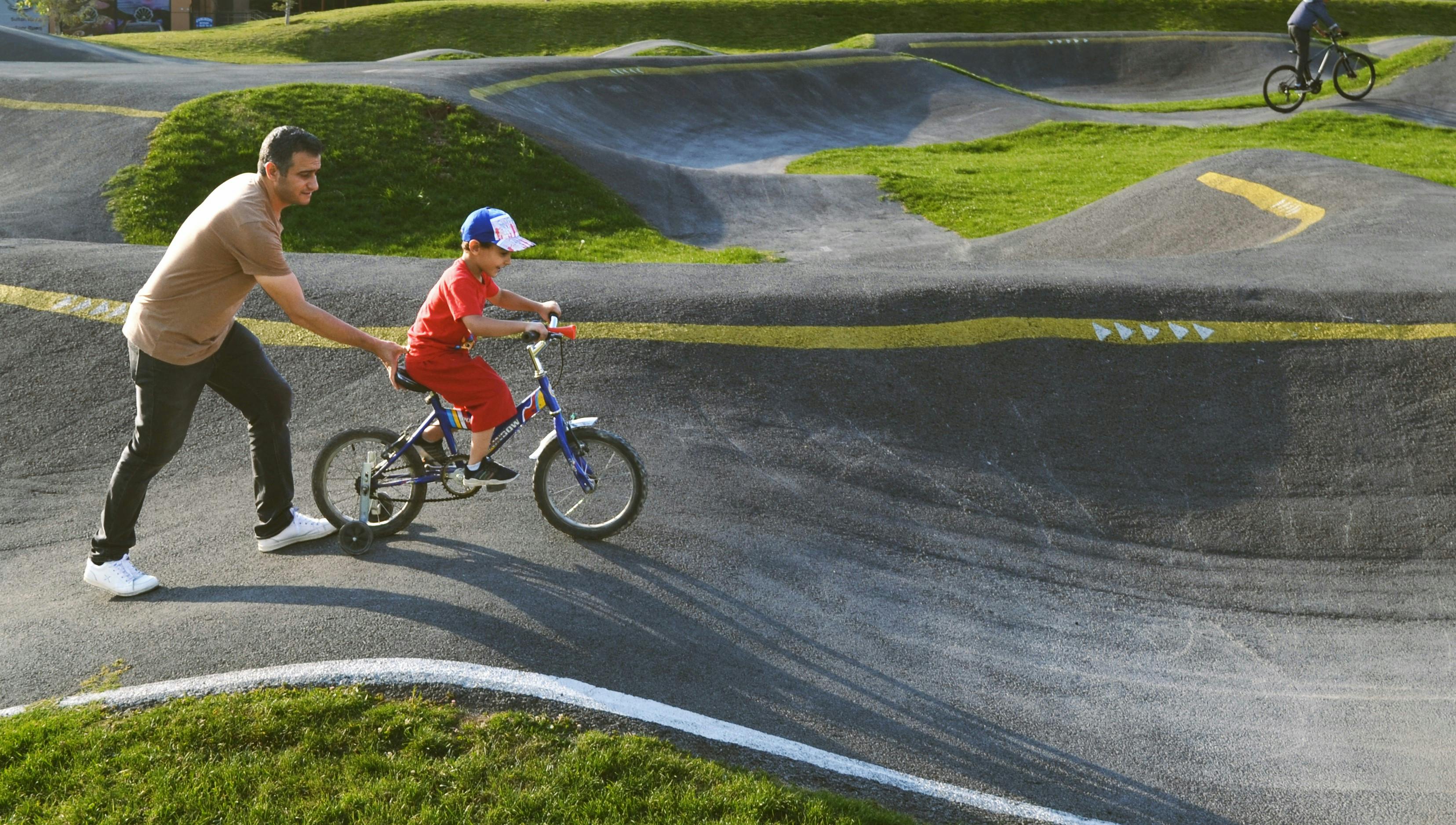 An adult helping a young child ride a small bike on a pump track, with another rider on a BMX in the background